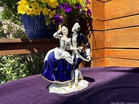 Porcelain figurine of a dancing couple outdoors on a purple cloth, showcasing the front and detail of painting and gold trim.