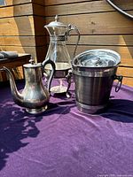 Photo showing the three items on a purple cloth surface outside, highlighting the shapes and details of each piece including the carafe, coffee pot, and ice bucket.