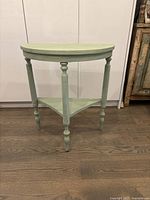 Front view of the half-moon shaped green wood accent table showing the turned legs and triangular lower shelf.