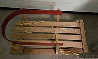 Overall view of wooden baby sled showing slatted seat, red curved seat back, yellow pull rope and runners