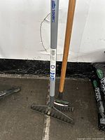 Two garden rakes standing on a textured black floor against a wall, one with a wooden handle and one with a grey handle.