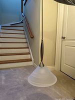 Modern floor lamp with frosted glass conical shade, metal pole with decorative midsection, standing by staircase and white door.