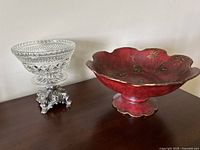 Pair of 2 pedestal bowls on wooden surface against white wall, showing clear cut glass bowl with silver metal ornate pedestal base and red scalloped rim bowl with floral and bird decoration.