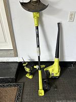 Front view of three Ryobi cordless garden tools and charger arranged upright against wall.