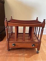 Full view of the cherry wood magazine rack featuring vertical slats, drawer with two knobs, and caster wheels on the legs.
