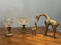 Full view of all four items together on wooden surface: crystal pedestal bowl, small crystal bowl, large and small brass giraffes