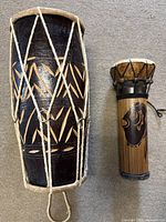 Front view of two drums, one large carved wooden body drum with natural skin head and white rope tuning, and one smaller drum with decorative body and black tuning rope.