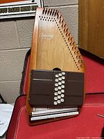 Front view of autoharp body standing upright on red case lining, showing chord bars, strings, tuning pins and logo