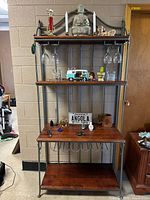 Full view of bakers rack styled bar showing metal frame and four wood shelves with various decorative items placed on shelves, and wine glasses hanging below second shelf.