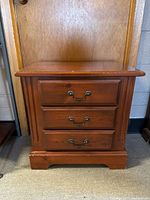 Front view of a medium brown wooden bedside table with three drawers, showing metal handles and signs of wear.
