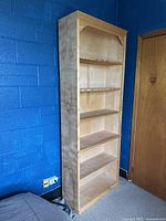 Front and side view showing the tall blonde wooden bookcase with five evenly spaced shelves and decorative trim on top edges.