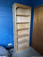 Full view of the blonde wood bookcase with five shelves against a blue wall and brown door.