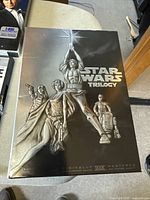 Front cover of the Star Wars Trilogy 4 DVD set showing molded artwork of main characters including Luke Skywalker with lightsaber, Princess Leia, Han Solo, C-3PO and R2-D2.