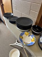 Plates and bowls in black matte finish with one decorative bowl, and some white cooking utensils in front.