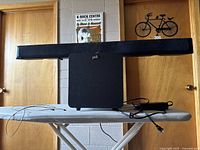 Front view of Polk Audio soundbar resting on wireless subwoofer with power supply and cords on ironing board.