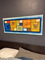 Framed rectangular painting with multiple Taino style blocks in colors of blue, orange, yellow, white, hanging on a grey wall above a bed with pillows.