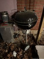 Full view of black Weber kettle grill with lid on tripod cart next to brick wall and fallen leaves.