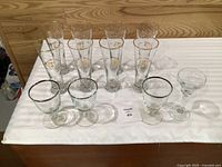 Full set of 12 glass pieces arranged: eight tall Olympic motif beer glasses, three silver-rim goblets, one coupe