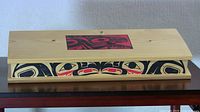 Photo of the closed cedar box showing the top with red and black Haida design centered on the lid and the overall shape.