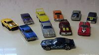 Photo showing all eleven Hot Wheels cars arranged on a surface, displaying a variety of colors and styles including a prominent navy blue car with blue flame decals.