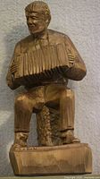 Full front view of wooden carved figure of a man seated with accordion, showing detailed carving and signature on base.