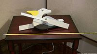 Full view of the entire pelican whirligig on a table showing its size relative to a measuring stick placed in front. The whirligig is painted white, black, gray with a yellow beak, and has wing pieces that are offset and attached with metal fasteners to move in opposite directions.