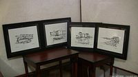 Four black framed prints are displayed resting on wooden tables against a light-colored fabric background. Each print shows a different black and white scenic illustration of Nova Scotia.