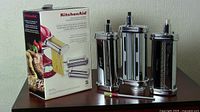 Three KitchenAid pasta maker attachments (roller, spaghetti cutter, fettuccine cutter) displayed next to original packaging, with clamp and handle visible.
