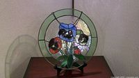 Photo showing a 13-inch round circular leaded glass sun catcher with colorful pansies floral design suspended by a black chain and displayed on a black stand.