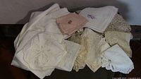 Photo showing an assortment of vintage linens including embroidered pillowcases, lace, and crochet pieces in white, beige, and pastel colors.