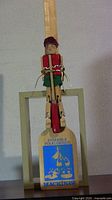 Front view of the wooden step dancing doll in traditional attire mounted on a paddle stage with Ensemble Folklorique Mackinaw branding