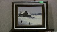 Framed acrylic painting of snow-covered barn scene in dark and silver frame against wall