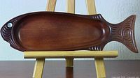 Overall view of the fish-shaped wooden dish with Tiki carvings on an easel showing the full shape and size.