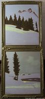 Photo showing two framed winter landscape pastel paintings stacked vertically on a table. Both with snowy scenes, pine trees, and mountains or water background.