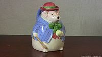 Front view of ceramic pig teapot with blue jacket and red hat, showing facial details and base.