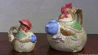 Front left view of rooster creamer and teapot, both holding blue ceramic cups, showing textured ceramic detail and coloration