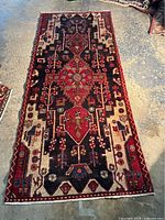 Full view of the rectangular rug on a concrete floor showing the geometric and floral medallion pattern in red, black, and cream colors.