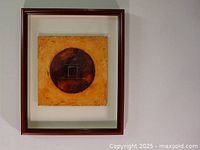 Front view of the framed abstract artwork showing a circular dark design on a mustard yellow background within a wooden frame.