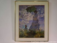Front view of framed art print depicting a woman holding an umbrella next to a child in a field with cloudy sky background, gold-tone frame and white matting.