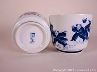 One porcelain tea cup shown alongside its underside with blue maker's mark visible. Blue illustrations of children and butterflies on the outside surface.
