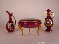 Front view of the makeup vanity set including the covered bowl and two small pitchers with gold handles and ornate hand-painted scenes.