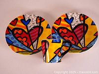 Top view showing one espresso cup and two saucers with colorful Romero Britto heart motif design.