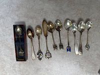 Overview photo showing all 10 silver plated souvenir spoons arranged on a flat surface. Some spoons display enamel portraits, others embossed designs.