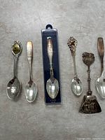 Photo showing 7 silver plated souvenir spoons with various Canadian regional designs, some with enamel details and relief scenes, all laid out on a grey surface.