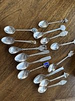 14 silver souvenir spoons arranged on wood surface showing varied decorative handles and bowl shapes