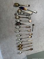 Full group of 16 decorative spoons and 1 butter knife laid out on a surface showing various handle designs and shapes.