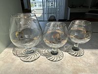 Three clear glass commemorative glasses with gold accents, arranged in sunlight showing the Olympic torch design and gold band rings.