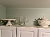 Photo showing three pieces: decorated white ceramic pedestal cake stand with tassels, clear glass pedestal bowl, white ceramic swirl-patterned bowl with lid.