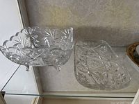 Crystal bowl and platter on a glass shelf, showing clear ornate cut crystal design with starburst patterns and scalloped edges on the bowl.