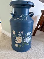 Full view of the blue metal milk can showing the floral decorations on the body and the lid with side handles.